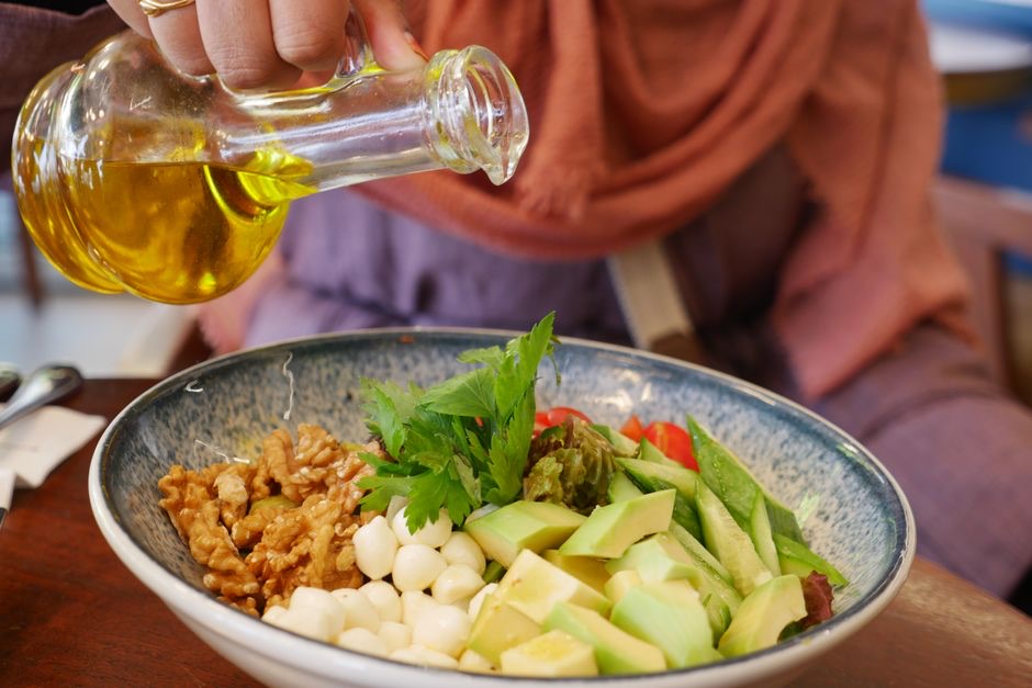 Main qui verse un filet d'huile d'olive sur un bol de salade aux noix, à l'avocat et au persil