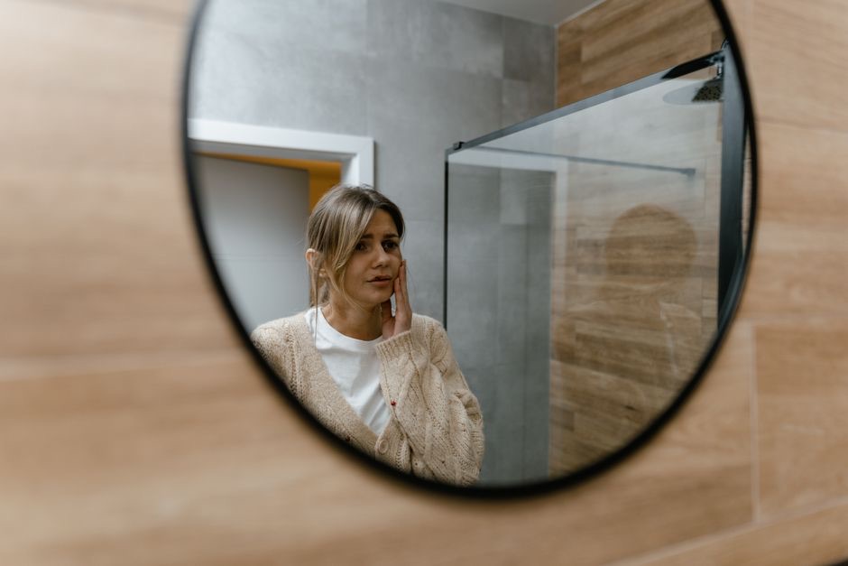 Reflet d'une femme en gilet beige dans un miroir rond de salle de bains, la main sur la joue