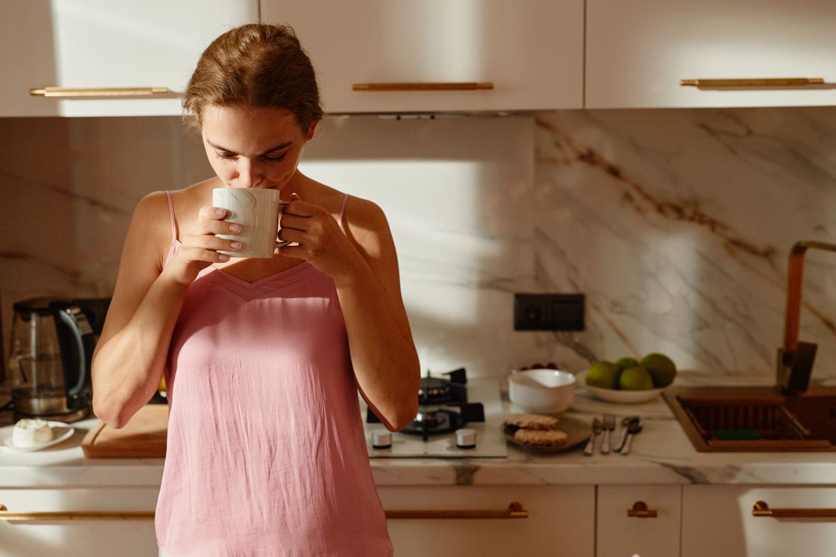 Femme en débardeur rose qui boit une tasse de café, debout dans sa cuisine baignée de soleil