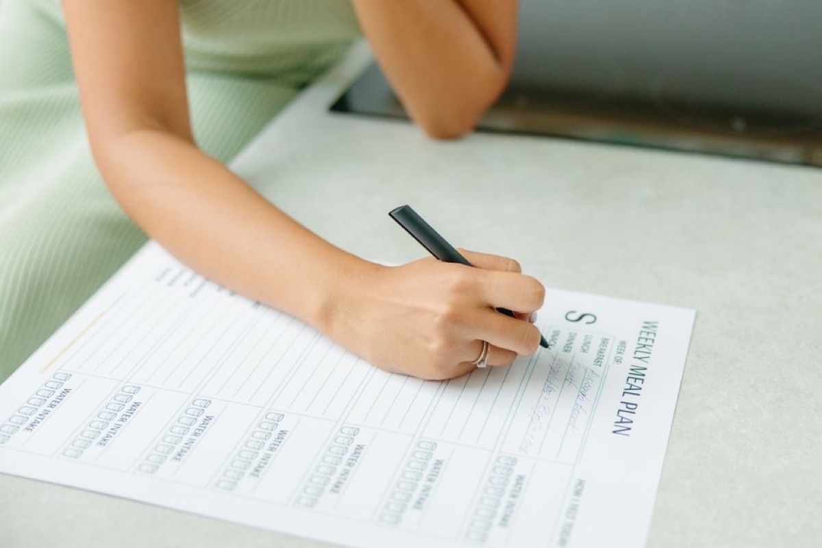 Femme qui note ses menus de la semaine sur un planning, chez elle, un stylo à la main