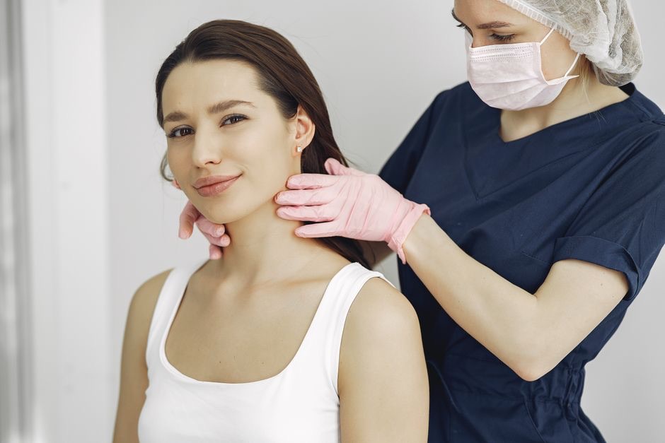 Soignante en blouse bleue et gants roses qui palpe le cou d'une jeune femme souriante